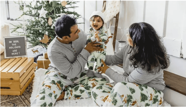 family of three sitting in front of a christmas tree. Mom and dad are both wearing cream colored pants with christmas trees and deer on them with a gray long sleeve shirt. Baby is wearing the same print in onesie style and a white hat. 